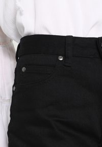 Mustat farkut slim fit -leikkauksella, viiden taskun designilla ja hopeansävyisillä yksityiskohdilla. Kankaan tekstuuri ja korkea vyötärötyyli näkyvät.
