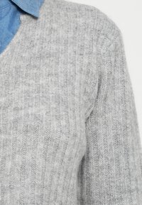 Svetlo sivi pleteni pulover z rebrasto teksturo in V-izrezom, prekrit z modro srajco s ovratnikom, ki prikazuje mehak material.