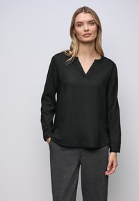 Blusa nera a maniche lunghe con scollatura a V, realizzata in tessuto morbido, con un taglio rilassato e tasche laterali, abbinata a pantaloni grigi testurizzati.