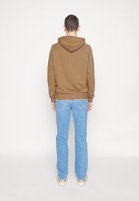 Sudadera marrón con un corte relajado, presenta una capucha con cordón y puños acanalados. Combinada con jeans rectos de color azul claro y zapatillas beige.