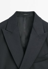 Blazer nero su misura con rever affilati, tessuto di lana strutturato, leggero lucido e taschino sul petto. Etichetta con la scritta "FABBRICATO IN PORTOGALLO."