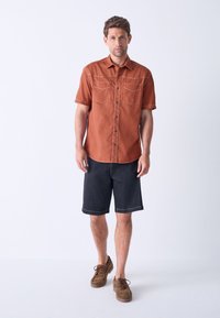 Short-sleeve, rust-colored button-up shirt with two chest pockets, paired with dark denim shorts and brown lace-up shoes.