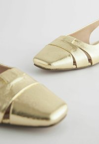 Złote płaskie buty z ekoskóry o wyraźnym, kwadratowym nosku. Posiadają trzy krzyżujące się paski oraz gładką fakturę, osadzone na neutralnym tle.