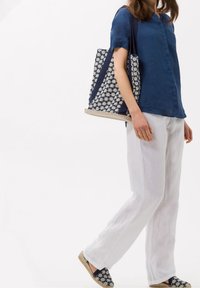 Donna che indossa pantaloni bianchi in lino, una camicia blu navy, porta una borsa a tracolla a fiori blu e bianchi e indossa scarpe slip-on floreali coordinate.
