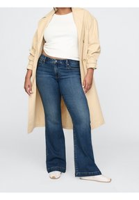 Kvinna som bär en beige trenchcoat, vit kortärmad topp, mörkblå utsvängda jeans och vita sneakers, stående mot en grå bakgrund.