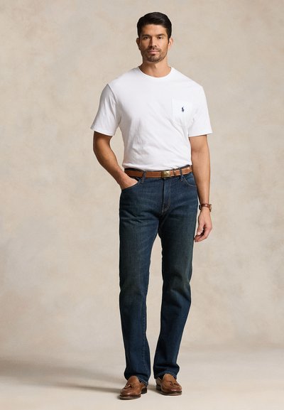 Mand iført hvid kortærmet T-shirt med lomme, mørkeblå jeans med brun bælte, ur og brune læder loafers, stående mod en ensfarvet baggrund.
