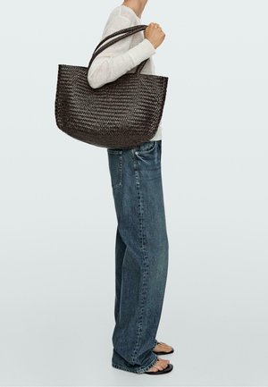 Persona che indossa jeans blu larghi e top bianco, tiene una grande borsa scura intrecciata sulla spalla.