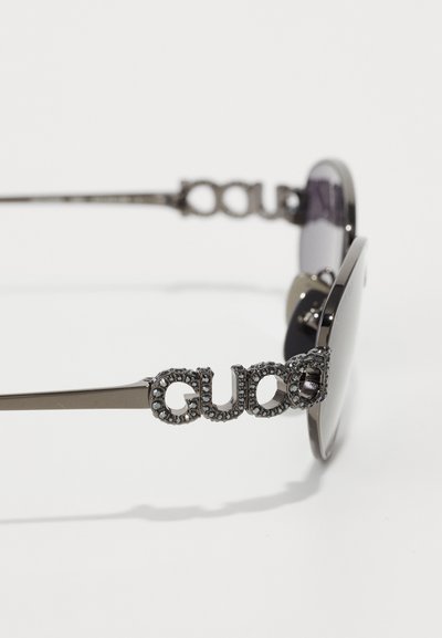 Naočale za sunce s tamnim lećama; metalni okvir srebrne boje; sadrži 'GUCCI' logo ukrašen rhinestones-ima na stablji.