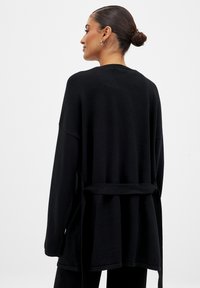 Svart långärmad cardigan med löst fall, sidslitsar och bindebälte vid midjan. Mjuk textur och minimalistisk design.