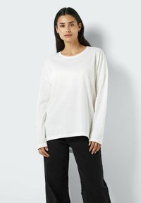Bijela dugohla t-shirt od laganog materijala s okruglim izrezom, s opuštenim krojem i spuštenim ramenima. Uparena s crnim hlačama.