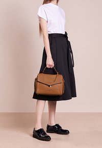 Sac à main en cuir texturé marron avec une anse supérieure, de forme rectangulaire et une fermeture magnétique. La modèle porte un t-shirt blanc et une jupe noire.