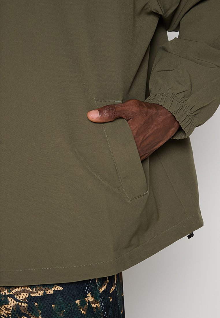 Chaqueta verde olivo con puños elásticos, que presenta un bolsillo lateral y una textura suave y lisa. Llevada sobre una tela con estampados, mostrando un diseño informal.