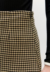 Schwarz-beige karierte Rock mit tailliertem Schnitt, glatter Textur und seitlicher Naht. Darüber ein schwarzes geripptes Oberteil mit langen Ärmeln.