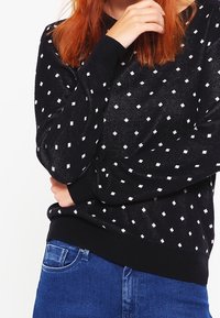 Czarny sweter z białym wzorem diamentu, długie rękawy, ściągacze na mankietach i u dołu. Noszony z niebieskimi dżinsami, prezentowany na białym tle.