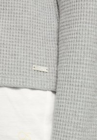 Helles graues, strukturiertes Sweatshirt mit Waffelstrickmuster und subtiler metallischer Optik. Ausgestattet mit einem silbernen Etikett mit "QS"-Markenlogo.