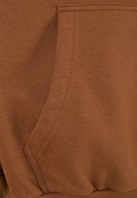 Gros plan d'un tissu marron avec des coutures visibles et une poche sur un vêtement, probablement un sweat-shirt ou un sweat à capuche.