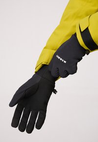 Gants isolés noirs avec une prise texturée et une sangle de poignet réglable, portés avec une veste jaune vif. Logo visible sur le poignet.