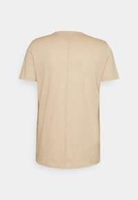 Vaaleanbeige lyhythihainen t-paita nähtynä takaa, jossa on pystysauman yksityiskohta keskellä, valkoista taustaa vasten.