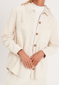 Krämfärgad kordjacka med krage, två framfickor och bruna knappar, med ribbad textur och avslappnad passform.