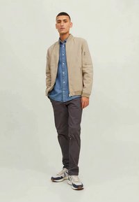 Light beige bomber jacket over a blue denim shirt, paired with dark gray pants and multi-color sneakers. Simple design with front zip.