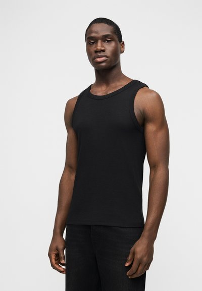 Άνδρας με σκούρο δέρμα, φοράει μαύρο αμάνικο τοπ και μαύρο παντελόνι, στέκεται μπροστά σε απλό γκρι φόντο.