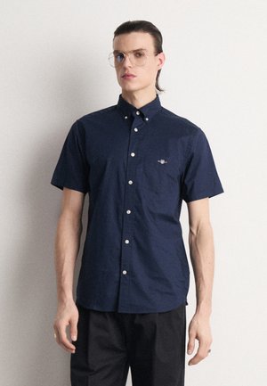 Chemise à manches courtes bleu foncé avec un col à bouton, dotée de boutons de couleur claire et d'un petit logo sur la poitrine. Tissu en coton lisse.