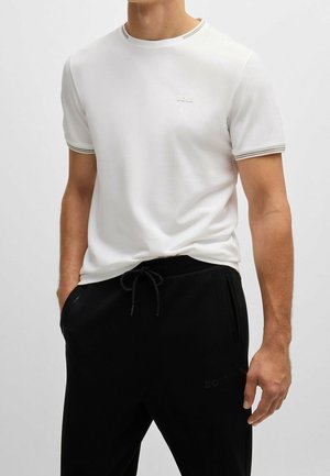 Mężczyzna w białym T-shircie z krótkim rękawem z logo „BOSS” oraz czarnych spodniach ze sznurkiem, stoi z rękami w kieszeniach.