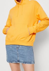 Gul huvtröja med framficka, snodd i huvan och ribbade ärmslut, tillsammans med en ljusblå denimkjol. Mjuk tygkvalitet och avslappnad passform.