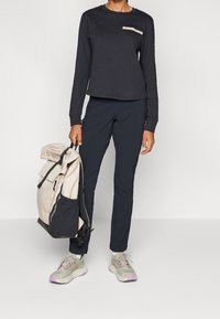 Black long-sleeve top with a white pocket detail, paired with dark pants and gray sneakers. A beige and black backpack is held in one hand.