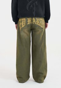 Olijfgroene wijde broek van denim, met grote gele tekst op de achterkant met een vervaagd effect. Achterzakken zichtbaar.