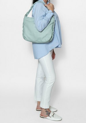 Lichtblauwe handtas met een gladde textuur en gevlochten hengsels, over de schouder gedragen, gecombineerd met een lichtblauwe top en witte broek.