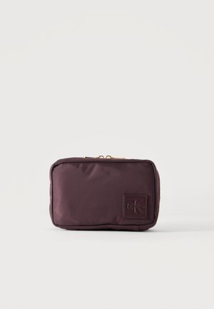 Lille rektangulær mørkebrun pung med lynlås og et Calvin Klein logomærke i nederste højre hjørne på en ensfarvet baggrund.
