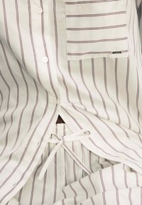 Nahaufnahme eines weißen Hemdes mit Knopfleiste und passender Hose mit dünnen braunen vertikalen Streifen, ausgestattet mit einer Brusttasche und einem Taillengürtel.