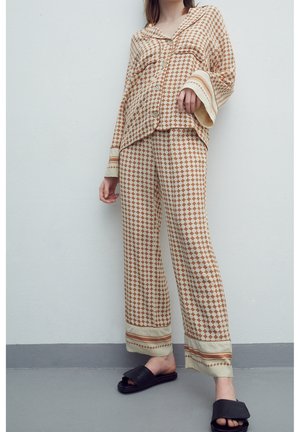 Femme portant une chemise à manches longues et un pantalon assortis à carreaux beige et marron, avec des sandales slides noires à bouts ouverts, debout contre un mur blanc.