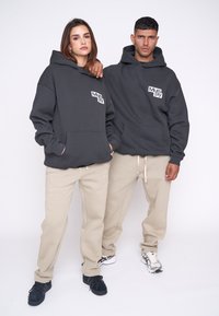 Sudaderas con capucha gris y bolsillos frontales, con un logo blanco, combinadas con pantalones de chándal beige. Personas que llevan zapatillas en blanco y negro están juntas.