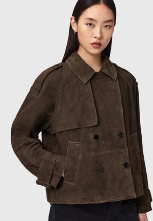 Jeune femme portant une veste croisée en suède brun foncé avec un large col et des détails à sangles sur les manches, sur un fond uni clair.