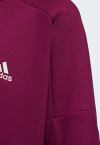 Närbild av en vinröd Adidas-tröja med ärm som visar logotypen i vitt broderad på det mjuka texturerade tyget.