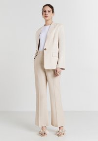 Woman standing in beige suit with tailored blazer, wide-leg pants, white top, rings, and floral-embellished open-toe heels on white background.