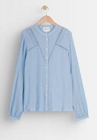 Lichtblauwe blouse met lange mouwen, knoopsluiting en een textuurstof. Heeft kantdetails langs de schouders en een ronde kraag.