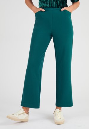 Pantalons larges en teal en tissu doux, dotés de poches latérales et d'une texture lisse. Portés avec des baskets blanches avec des accents beiges.