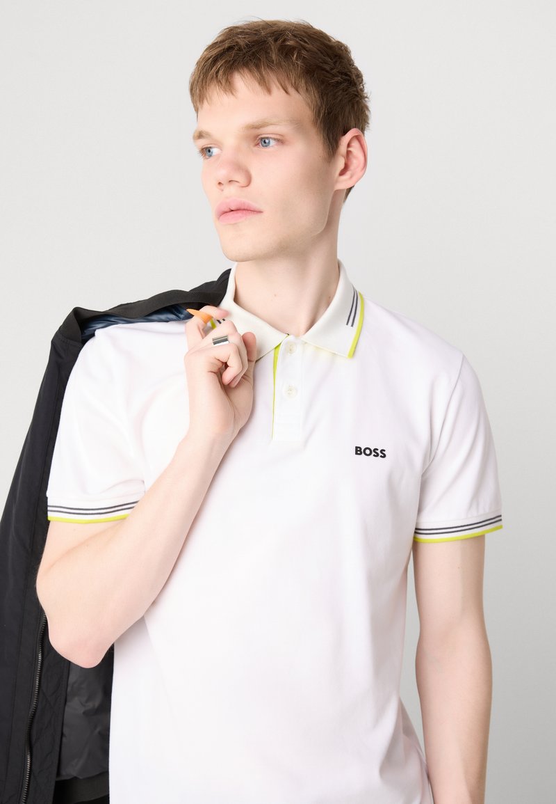 Biała koszulka polo z czarno-żółtym kołnierzykiem w paski, z logo "BOSS". Model trzyma czarną marynarkę na ramieniu.