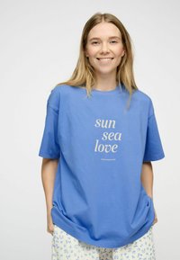 Sininen puuvillatrikoo t-paita lyhyillä hihoilla ja väljällä mitoituksella, jossa on vaaleasta väristä teksti "aurinko meri rakkaus". Yhdistettynä vaaleisiin kuviollisiin pöksyihin.
