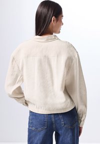 Krämfärgad, lätt jacka med avslappnad passform, som har en krage och elastisk midja, tillsammans med blå denimjeans.