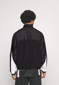 Man with curly hair wearing a black jacket with white stripes on sleeves, standing facing away against a plain light background.