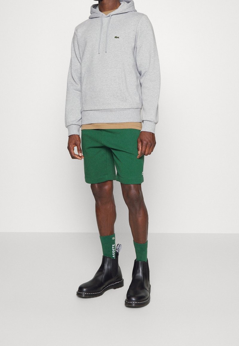 Uomo che indossa un hoodie grigio chiaro, una maglietta beige sottostante, pantaloni corti verdi, calzini verdi Lacoste e stivaletti neri in pelle, in piedi su un pavimento grigio.