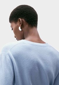 Jasnoniebieski sweter o miękkiej fakturze i okrągłym dekolcie. Drapowanie sięga do ramion, odsłaniając srebrną kolczyk w uchu.