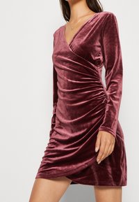 Sammetklänning i bärnstensröd, med V-hals, långa ärmar, rynkade detaljer på sidan och en kroppsnära silhuett. Slät textur.