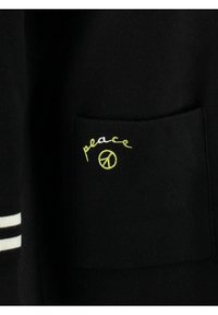 Tessuto nero con una tasca applicata. Ricamato "pace" in corsivo giallo e un dettaglio con simbolo della pace giallo. Strisce bianche sulle maniche.