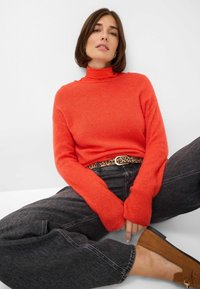 Helle orange Rollkragensweater aus weichem Material, kombiniert mit dunkelgrauen Jeans und einem Gürtel mit Leopardenmuster. Braune Wildlederschuhe runden den Look ab.