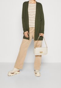 Cardigan lungo verde oliva sopra una maglietta a righe beige, abbinato a pantaloni beige a gamba larga e zoccoli chiari. Tiene in mano una borsa crema.